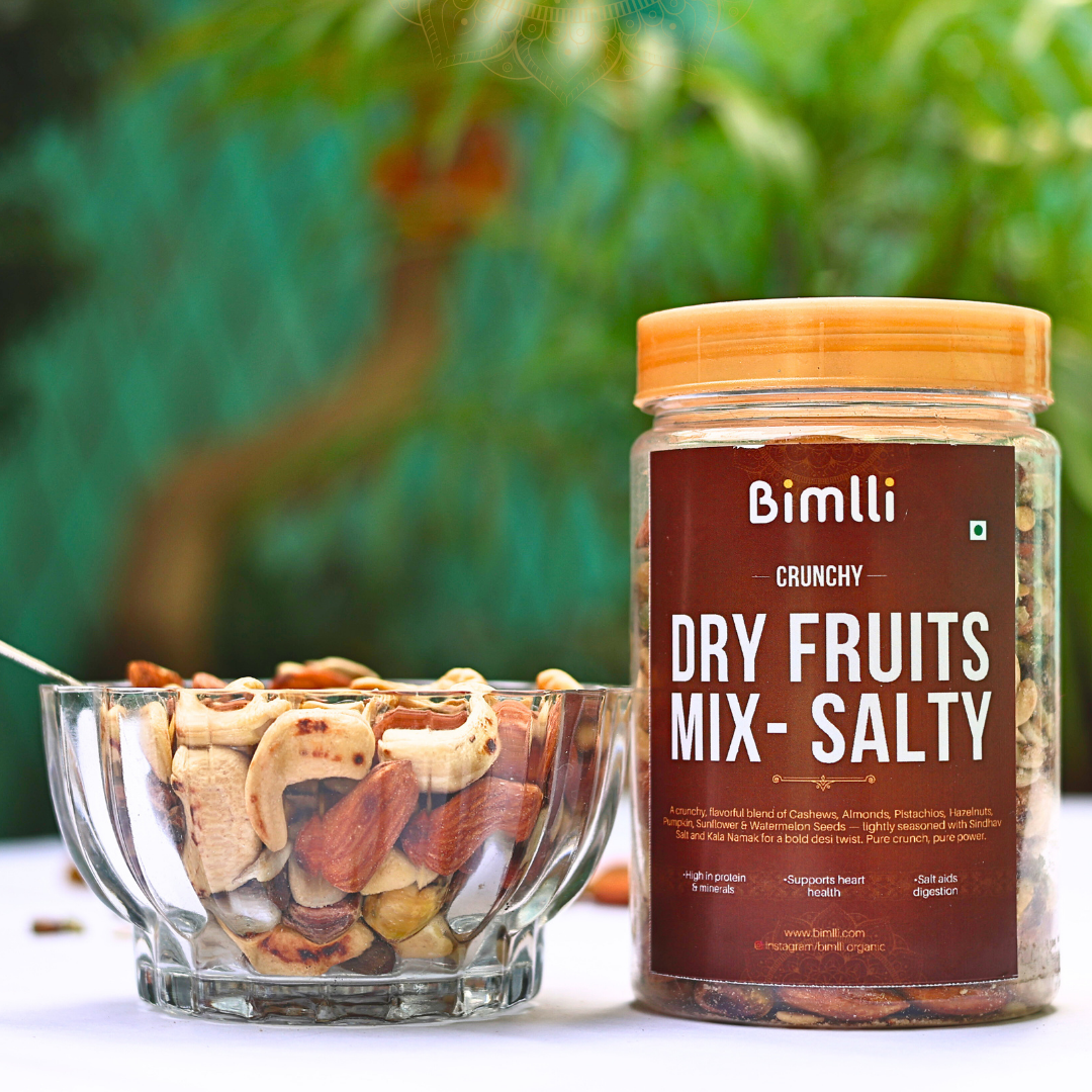 Jar of Bimlli Crunchy Dry Fruits Mix with a bowl of mixed nuts on a blurred green background