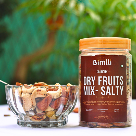 Jar of Bimlli Crunchy Dry Fruits Mix with a bowl of mixed nuts on a blurred green background